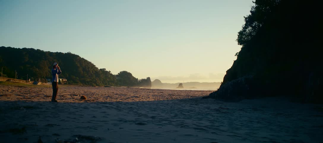 A lone man stands on a tranquil sandy beach and takes a photo of the beautiful coastal scenery during a golden sunrise