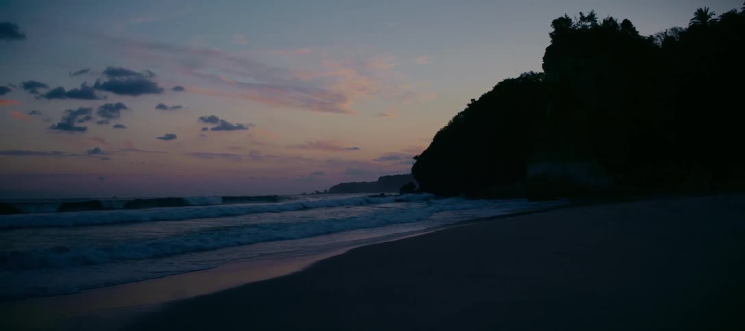 A tranquil beach at twilight where gentle waves wash ashore next to a silhouetted cliff