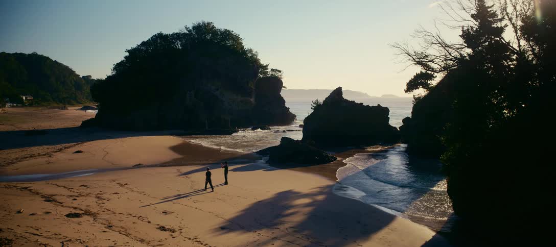 Two silhouetted men practice sword fighting on a serene sandy beach with large rock formations at sunrise