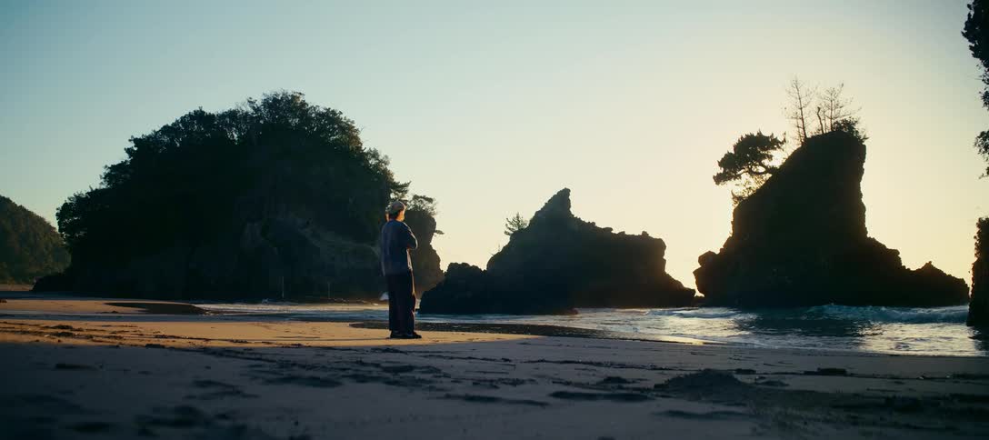 A solitary figure enjoys a peaceful walk on a rugged beach at sunrise, pausing to watch the waves crash against the silhouetted rock formations
