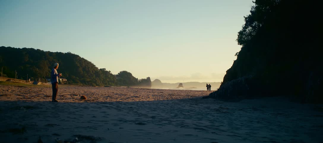 A person photographs other beachgoers from a distance on a vast, sandy shore during a tranquil golden hour sunrise