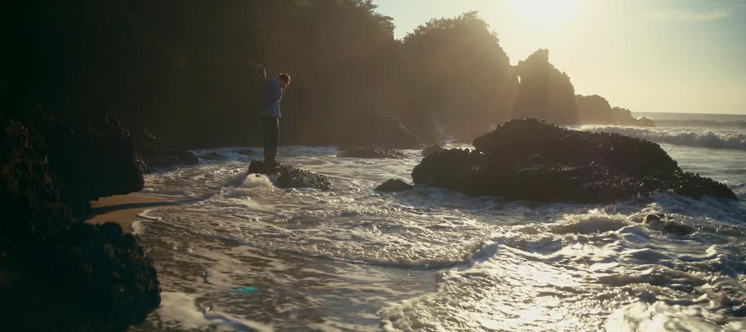 A photographer stands on a rock at a hazy, sun-drenched beach, capturing the slow-motion crash of waves against the shore