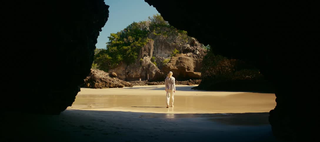 A woman emerges from a dark cave and walks barefoot onto a secluded, sun-drenched beach surrounded by rocky cliffs