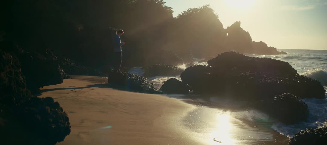 A solitary figure stands on a rock at a secluded beach, watching the ocean waves during a beautiful golden sunrise