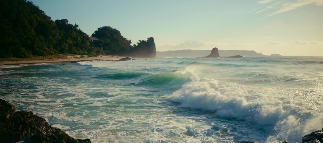 A scenic wide shot captures gentle ocean waves rolling onto a rocky coastline in slow motion during the golden hour