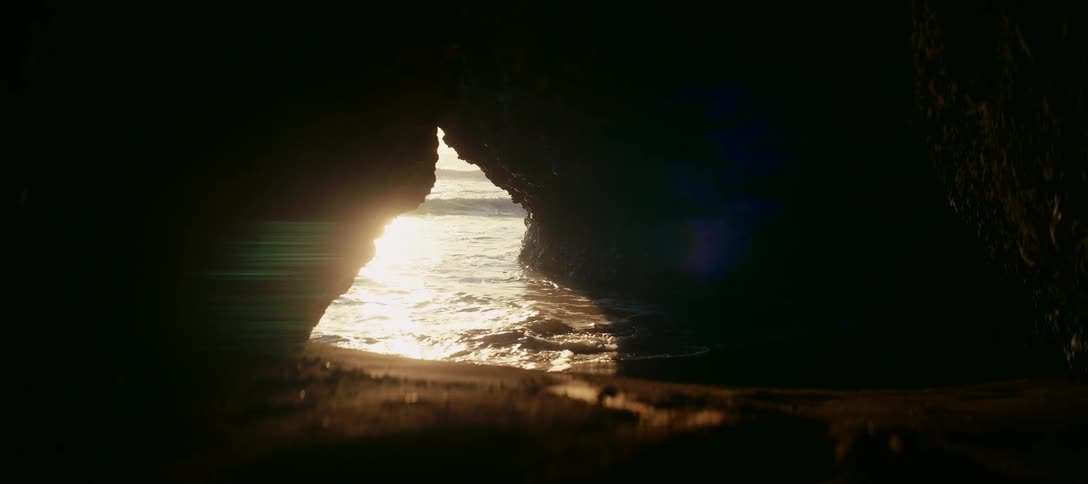A tranquil view from inside a dark sea cave shows the golden sunset reflecting on the slow-motion ocean waves washing ashore