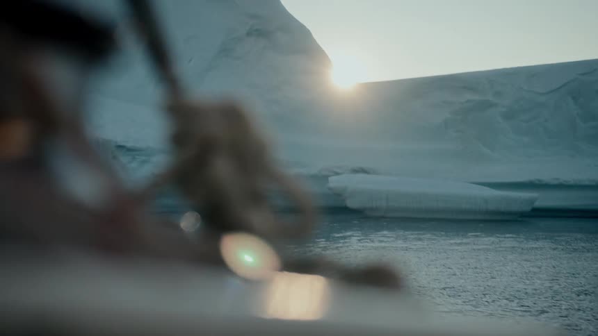 A boat sails through the Arctic waters past a massive iceberg as the sun sets, casting a warm glow and lens flare