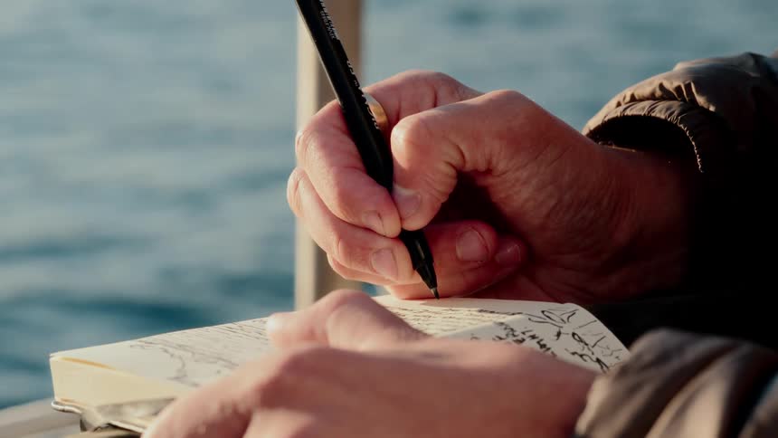 A close-up of a person's hands sketching in a journal with the ocean in the background