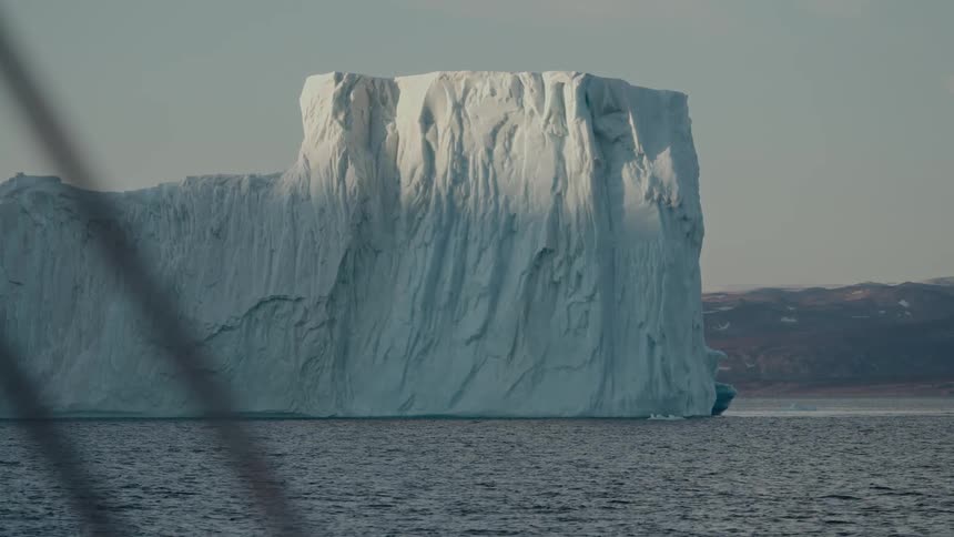 A massive iceberg floats in the calm ocean under a hazy sky, illuminated by the sun
