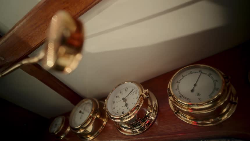 A low angle shot of beautiful brass nautical instruments, including a clock and thermometer, gently rocking inside a vintage ship's cabin
