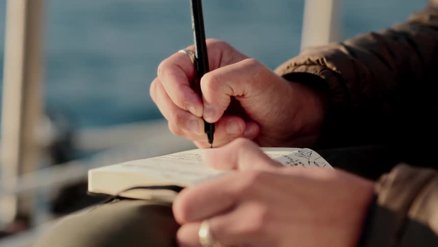 A slow-motion close-up of a person's hands journaling in a notebook with the ocean in the background