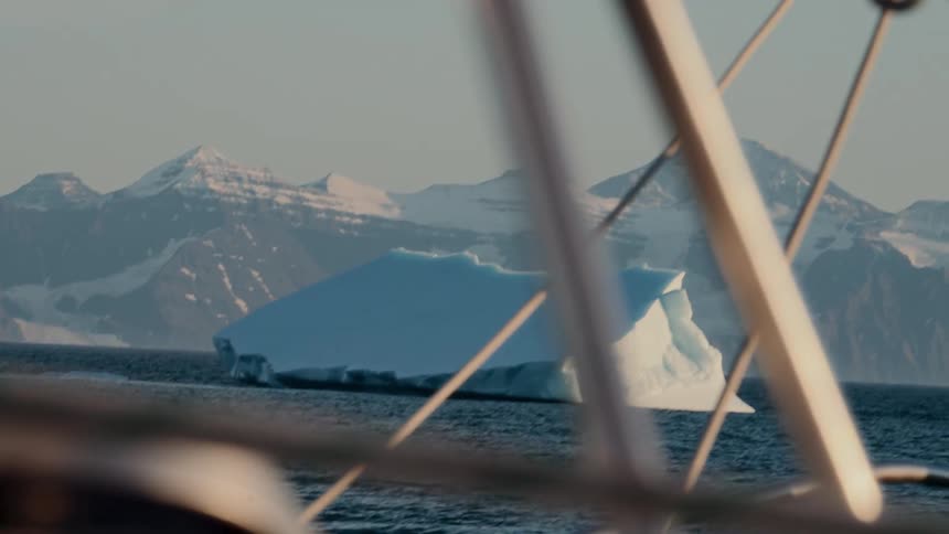 A serene view of a massive iceberg floating in the ocean with snow-covered mountains in the background, as seen from the deck of a sailing boat
