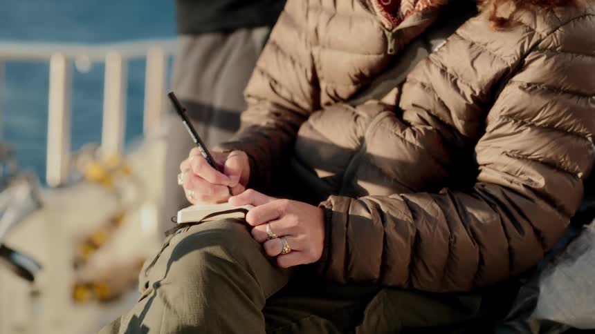 A woman in a warm jacket sits on the deck of a boat and writes in her journal as the sun shines
