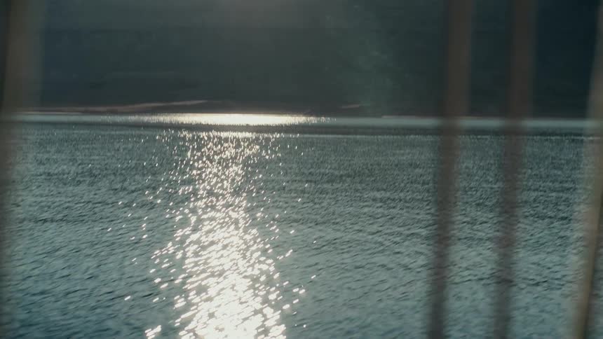 Peaceful view through reeds of the sun reflecting and sparkling on a calm lake with mountains in the distance