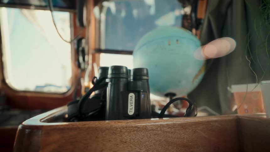 A close-up shot of binoculars and a globe sitting on a wooden desk inside a gently rocking boat cabin
