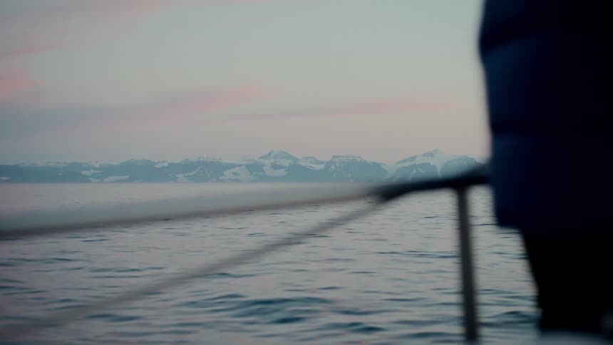 A view from a boat sailing on a calm ocean towards a snow-covered mountain range at dusk
