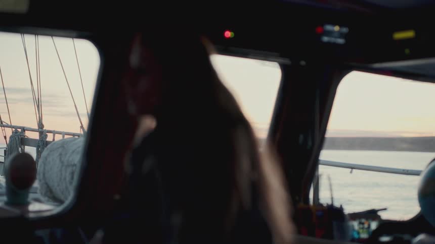 A silhouetted woman with long hair pilots a boat from the wheelhouse during a beautiful sunset over the ocean