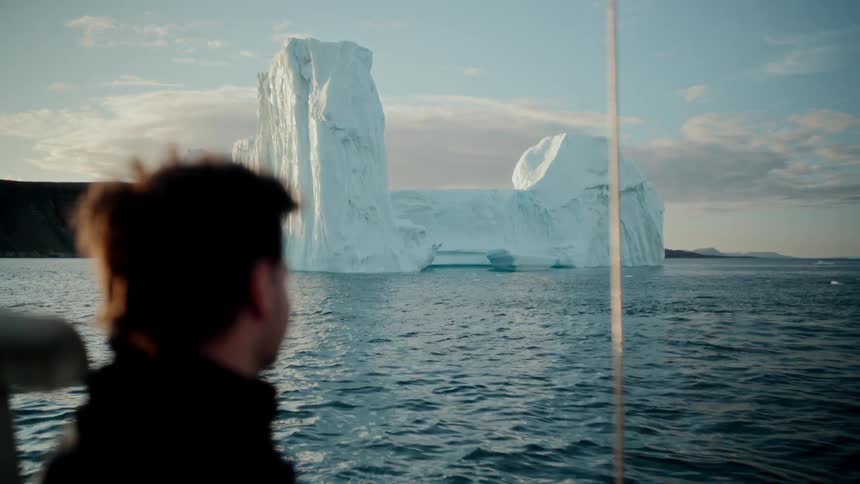 A man on a boat journey admires a colossal iceberg floating in the arctic ocean during a scenic golden hour