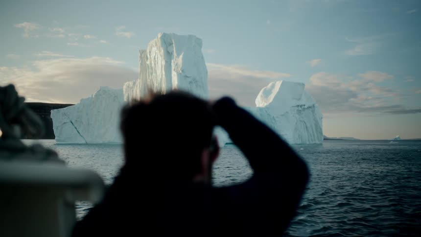 A silhouetted traveler on a boat expedition photographs a majestic iceberg floating in the arctic ocean