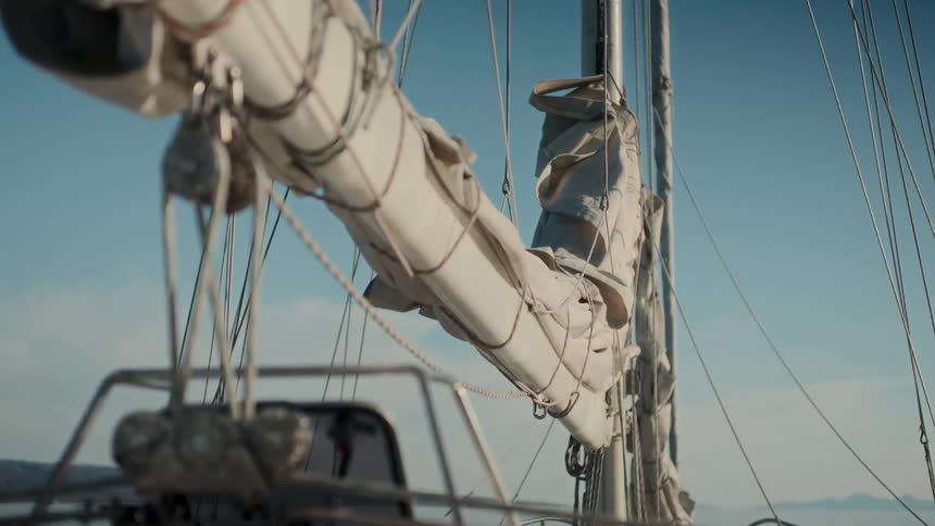 A close-up of a furled mainsail on a gently rocking sailboat under a clear blue sky