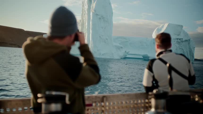 Two men on a boat photograph a massive iceberg floating in the calm arctic ocean during a golden sunset