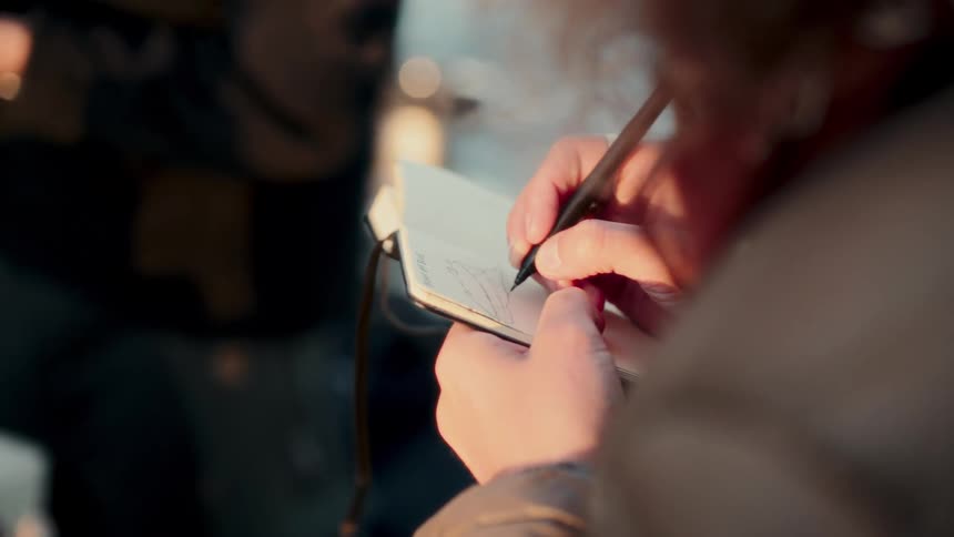 An artist carefully sketches a simple line drawing in their pocket-sized notebook with a fine-tipped pen