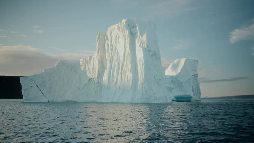 A colossal iceberg floats serenely in the vast ocean, illuminated by the warm light of the sun