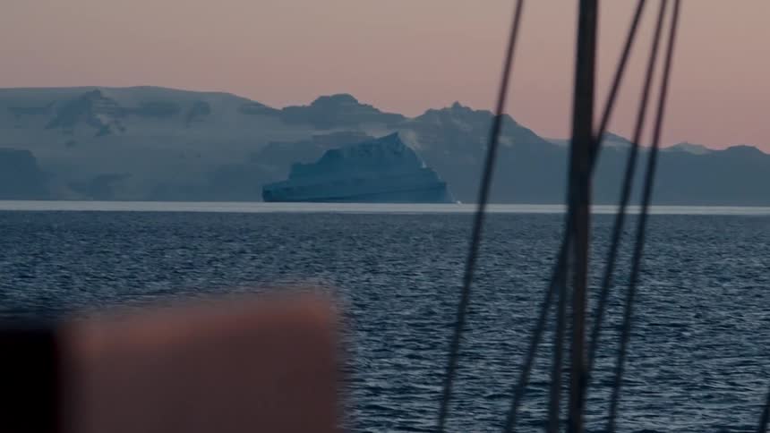 A serene view from a sailboat captures a massive iceberg floating in the arctic ocean with snow-covered mountains in the distance at dusk