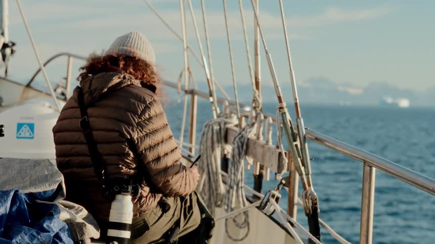 A photographer bundled in warm clothes sits on the deck of a sailboat, admiring the vast arctic ocean with distant icebergs and snowy mountains