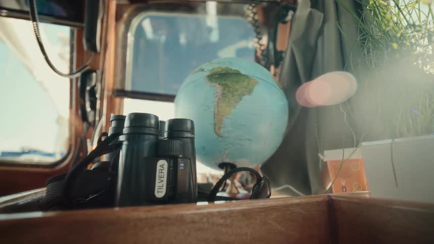 A slow push-in shot of binoculars and a globe resting inside a sunny boat cabin evokes a sense of wanderlust and adventure