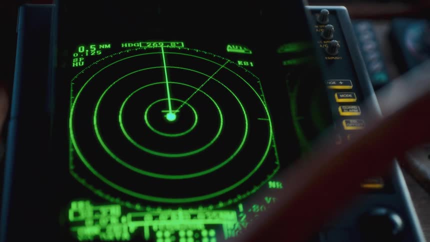 A captain's hand operates a green marine radar screen showing a sweeping beam on a ship's bridge