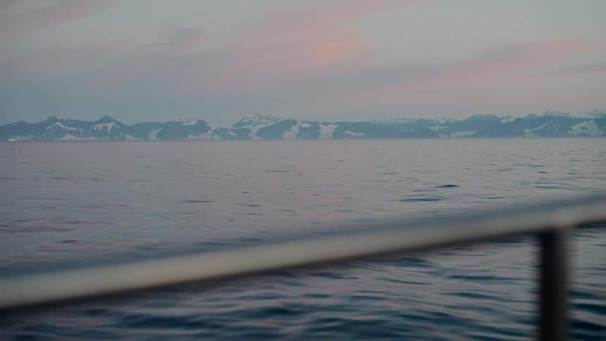 A serene view from a boat sailing across calm waters towards a distant, snow-covered mountain range under a pastel-colored sky at dusk