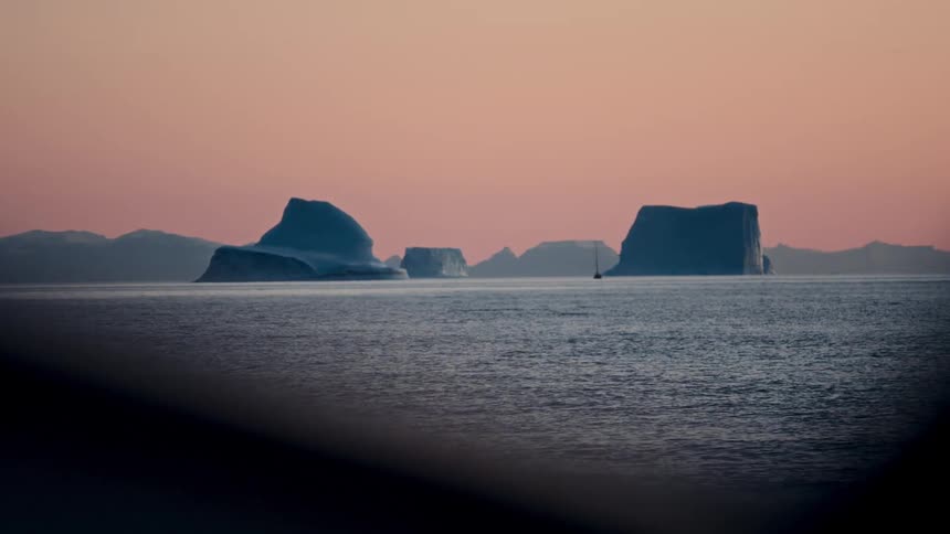 Majestic icebergs and a lone sailboat float on the calm arctic ocean against a beautiful pink sunset sky