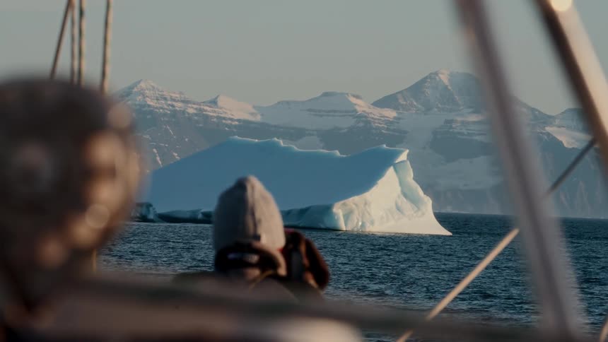 From the deck of a sailboat, an explorer gazes at a majestic iceberg floating in the arctic sea against a backdrop of snow-capped mountains