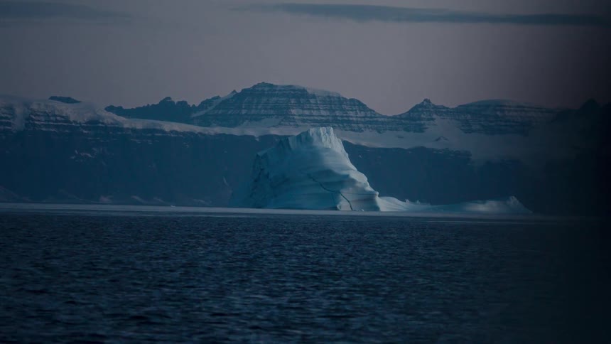 A majestic iceberg drifts through the dark arctic sea at twilight, with snow-covered mountains in the distance