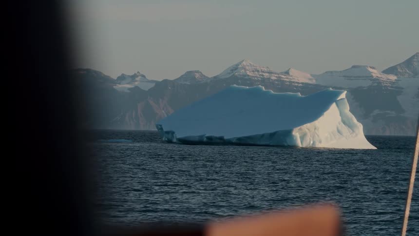 An iceberg drifts in the arctic ocean with snow-covered mountains in the background, as seen from a sailboat