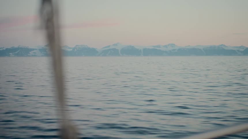A peaceful view from a sailboat shows the calm sea and a distant range of snow-capped mountains on the horizon at dusk