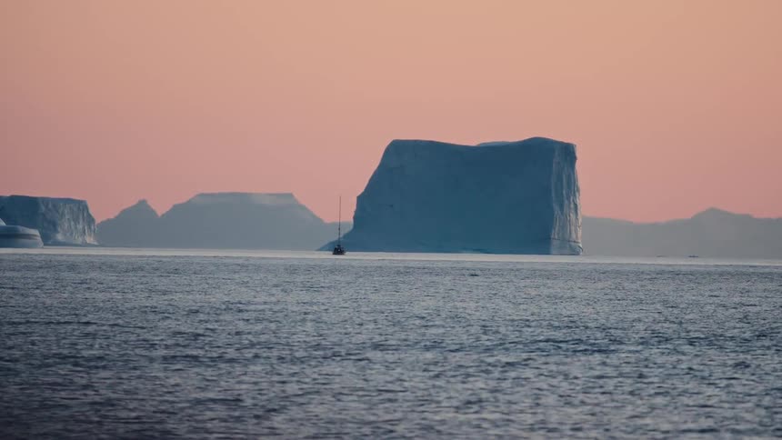 A lone sailboat navigates through massive icebergs floating in the arctic ocean during a beautiful pink sunset