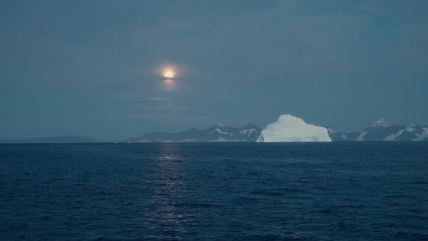 A bright full moon shines over a vast arctic ocean where a large iceberg floats near a snowy mountain range