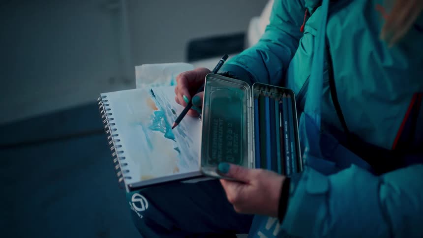 Close up of an artist in a blue winter jacket sketching a landscape with colored pencils in a notebook