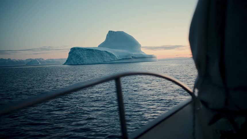 A sailboat glides through calm arctic seas, offering a breathtaking view of a giant iceberg at dusk