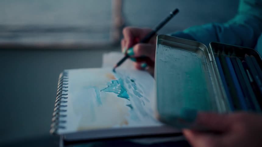 A close-up view of an artist with vibrant nail polish sketching a colorful landscape in a notepad with a pencil