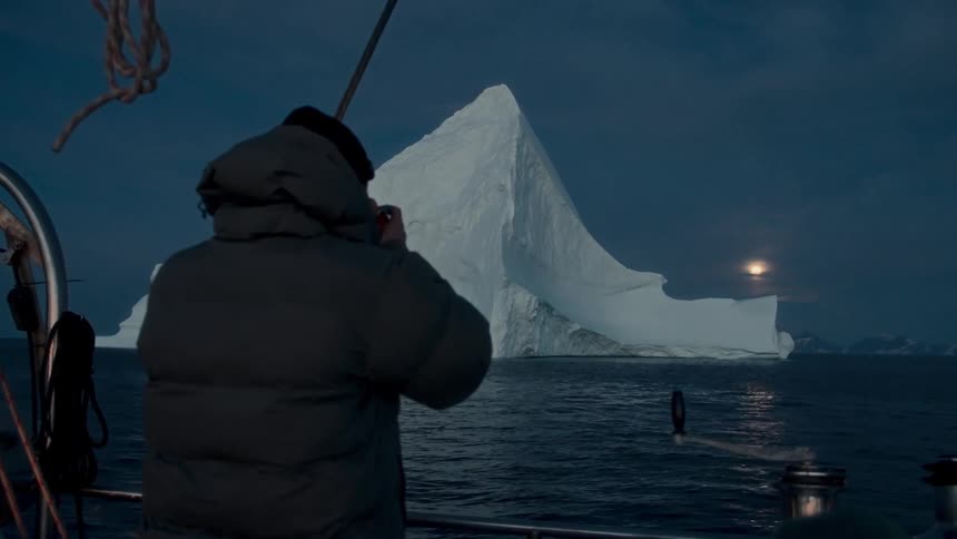 A traveler on a sailboat photographs a majestic iceberg as the full moon rises over the arctic sea at twilight