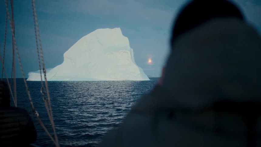 Adventurers on a sailboat watch and photograph a massive iceberg floating in the arctic ocean at dusk
