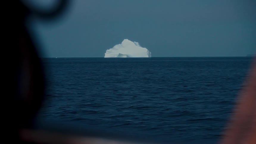 A solitary iceberg floats on the distant horizon across the vast, deep blue ocean as viewed from a boat