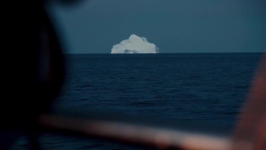A solitary iceberg drifts in the deep blue sea as seen from the railing of a ship's deck