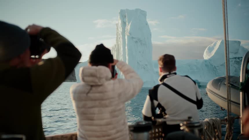 A group of friends on a sailboat take photos of a majestic iceberg floating in the calm arctic ocean at sunset