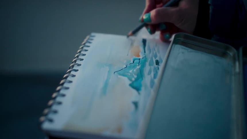 Close-up of an artist adding detail with a colored pencil to a watercolor landscape in a sketchbook