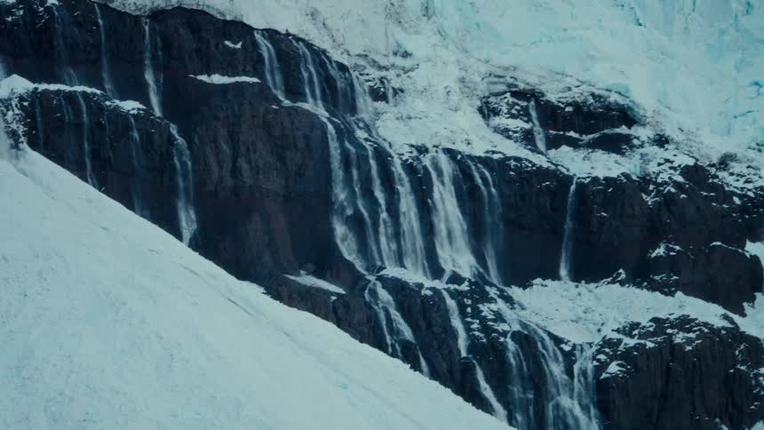 Waterfalls from a melting glacier cascade down a dark, rocky cliff face in a vast, snow-covered landscape