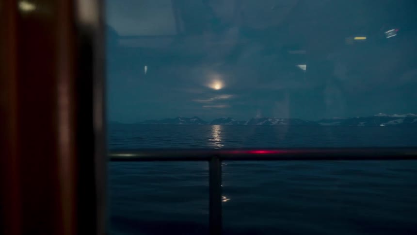 A serene view from a boat window shows the moon reflecting on the ocean with snowy mountains on the horizon